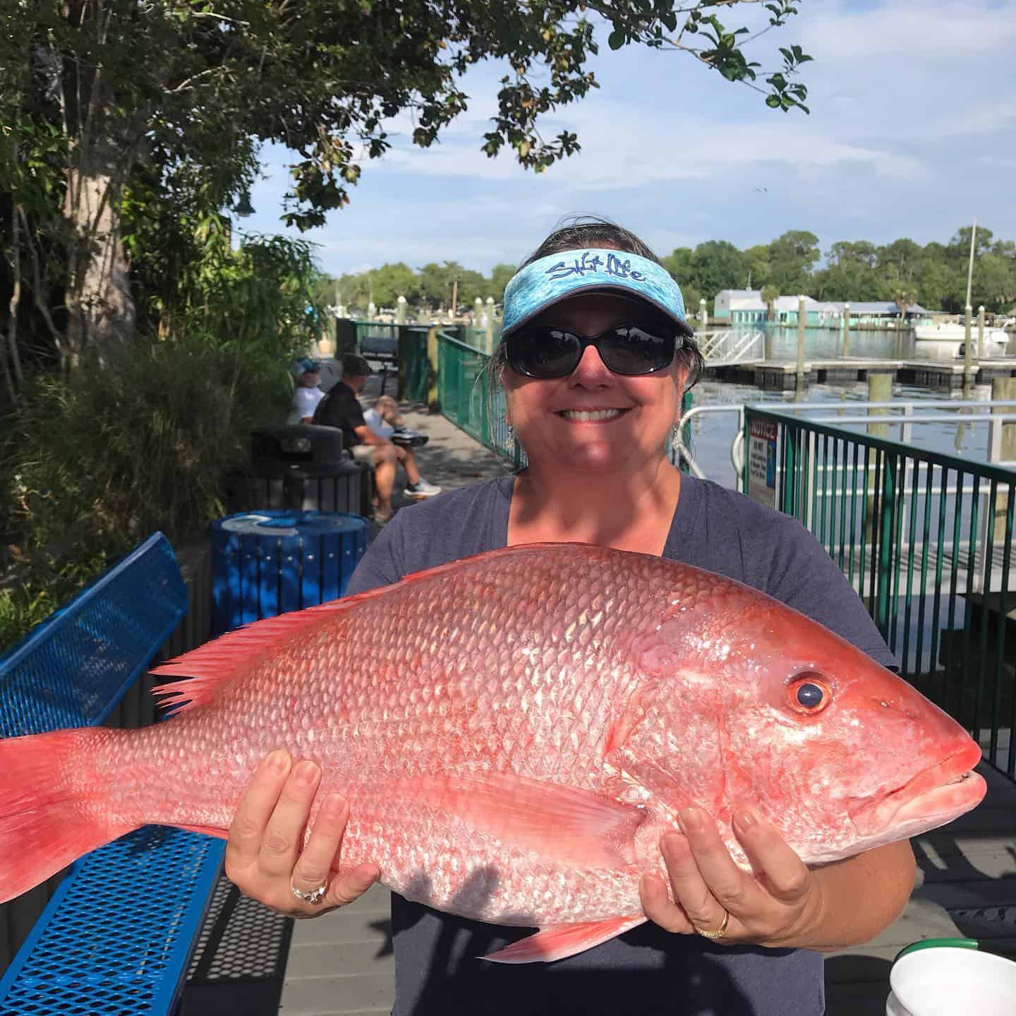 A smiling woman wearing sunglasses and a blue visor holds a large, freshly caught red snapper fish while standing on a dock with boats and greenery in the background.